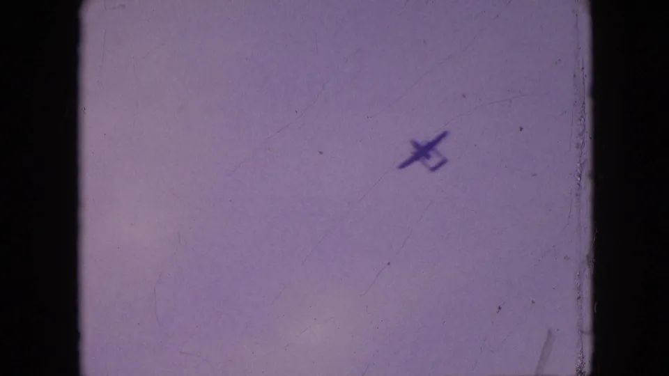A small propeller plane is silhouetted against the cloudy Philadelphia sky near the right side of the 8mm frame, with a roof edge partly visible below.