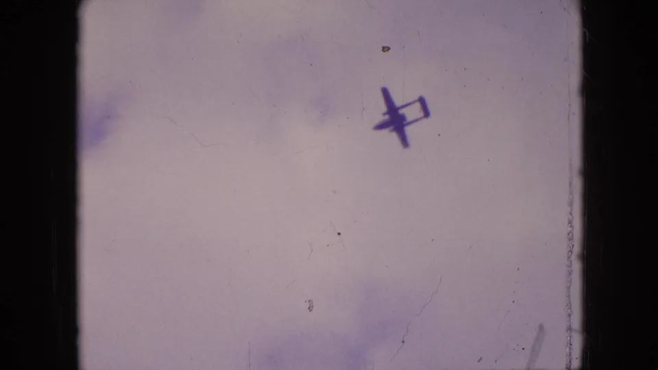 Dark aircraft silhouette angled across a faded sky in 1964 Philadelphia 8mm footage, with scratches and light color variation visible.