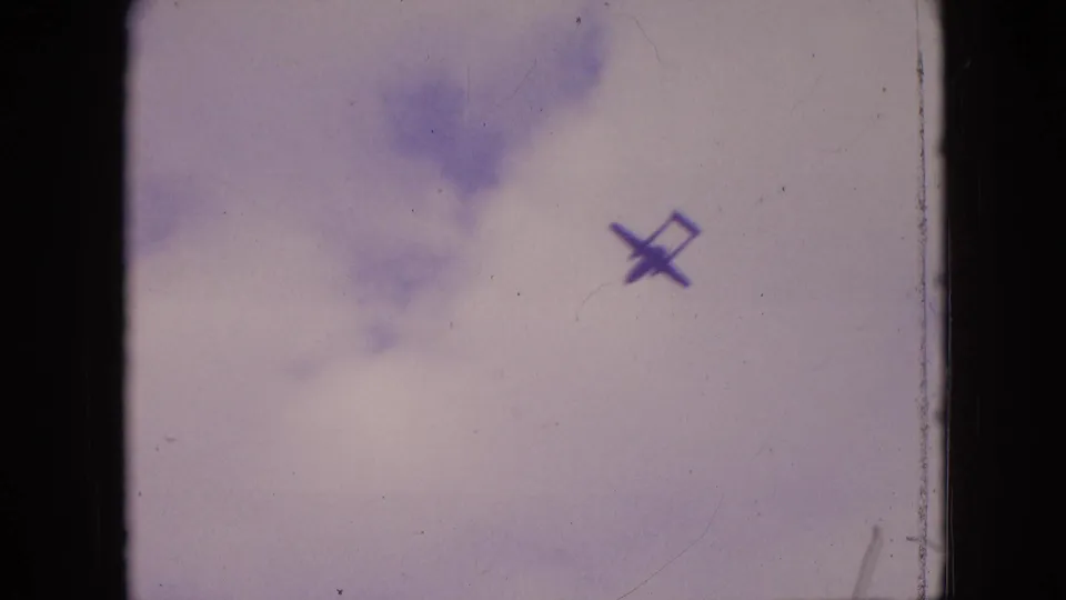 Small aircraft silhouette passing beneath soft cloud patches in a faded sky, shown in supplied 1964 Philadelphia home-movie footage.
