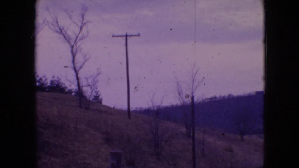 1964 Philadelphia-area 8mm view of a telephone pole centered on a grassy sloped hill with bare trees, distant low hills, and an overcast sky.