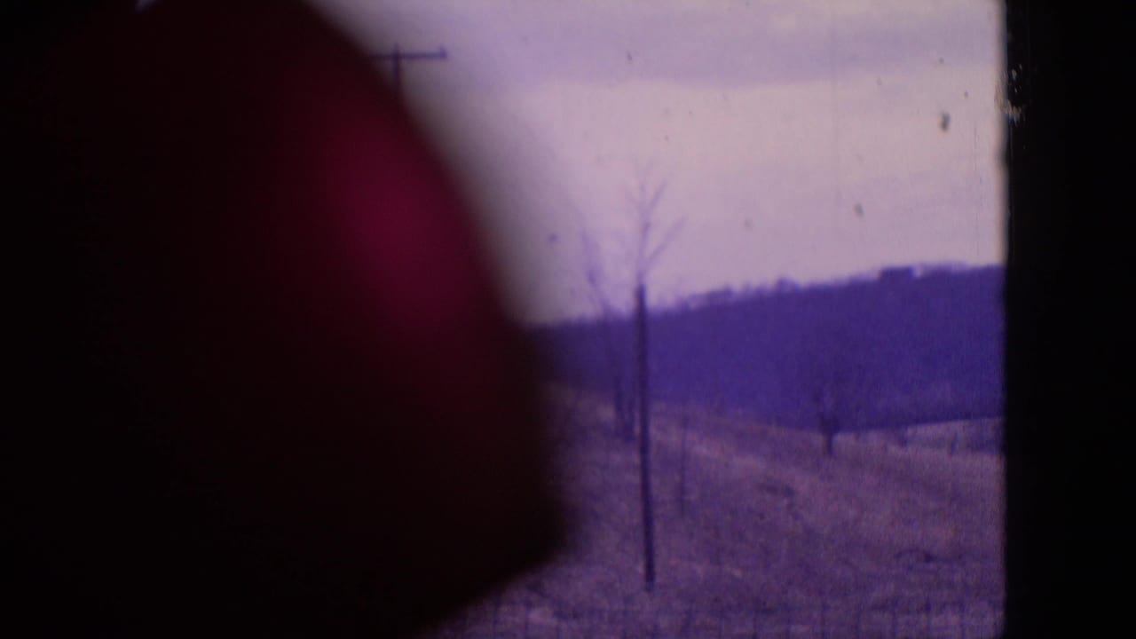 1964 Philadelphia-area 8mm frame showing a rural hillside with bare trees, utility poles, distant low hills, cloudy sky, and a dark rounded obstruction at left.