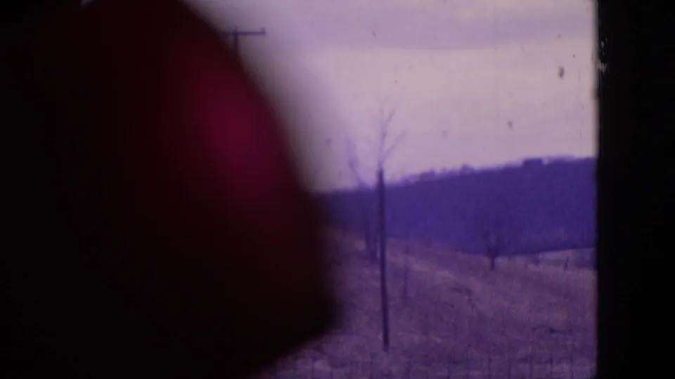 1964 Philadelphia-area 8mm frame showing a rural hillside with bare trees, utility poles, distant low hills, cloudy sky, and a dark rounded obstruction at left.