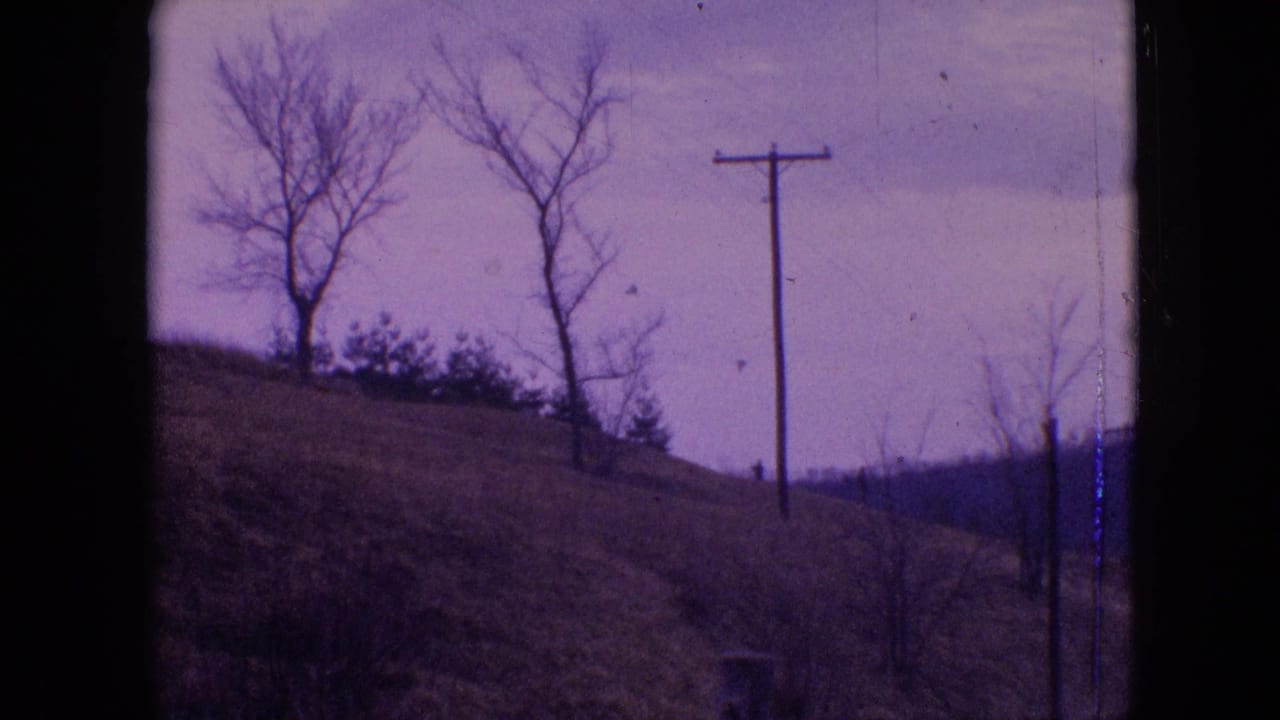 1964 Philadelphia-area home-movie frame with bare trees on a sloped hillside, a wooden utility pole, brush, cloudy sky, and dark vertical border from the film or viewpoint.