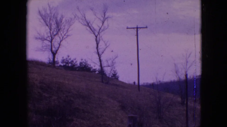 1964 Philadelphia-area home-movie frame with bare trees on a sloped hillside, a wooden utility pole, brush, cloudy sky, and dark vertical border from the film or viewpoint.