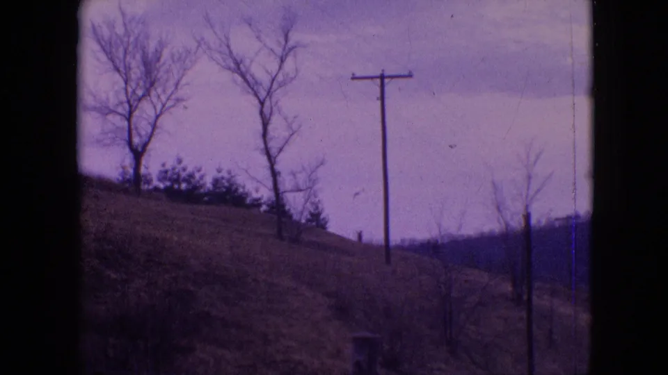 1964 Philadelphia-area 8mm frame showing a grassy hillside with bare trees, a utility pole, brush, distant hills, cloudy sky, and purple color cast from aged film.