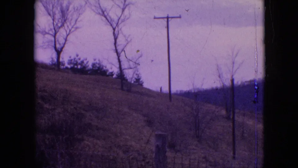 1964 Philadelphia-area rural landscape frame with a sloping hill, bare trees, wooden utility pole, brush, distant ridge line, cloudy sky, and visible 8mm edge wear.