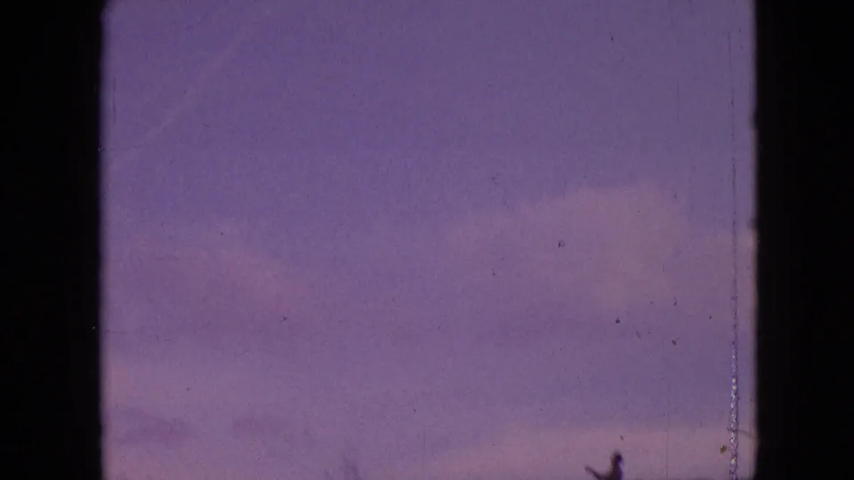 1964 Philadelphia home-movie frame with purple sky, low hillside, faint clouds, scratches, and a small silhouetted person standing near the lower right.