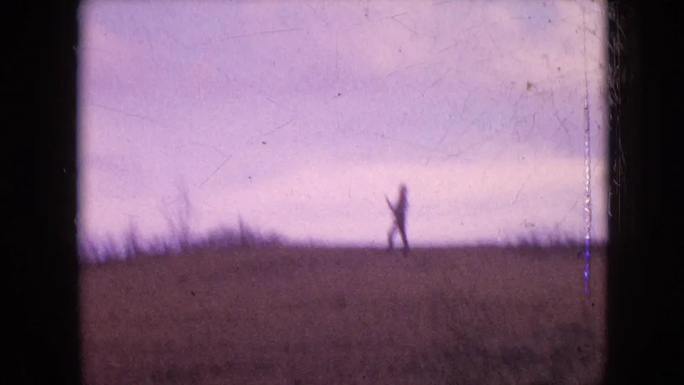 A distant silhouetted figure, possibly managing a kite line, stands on the crest of an open field in this faded 1960s 8mm frame.