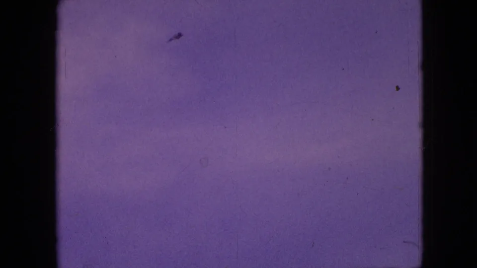 A small dark shape, possibly a kite, appears against the purple-blue sky above the open field in this scratched 8mm home-movie frame.