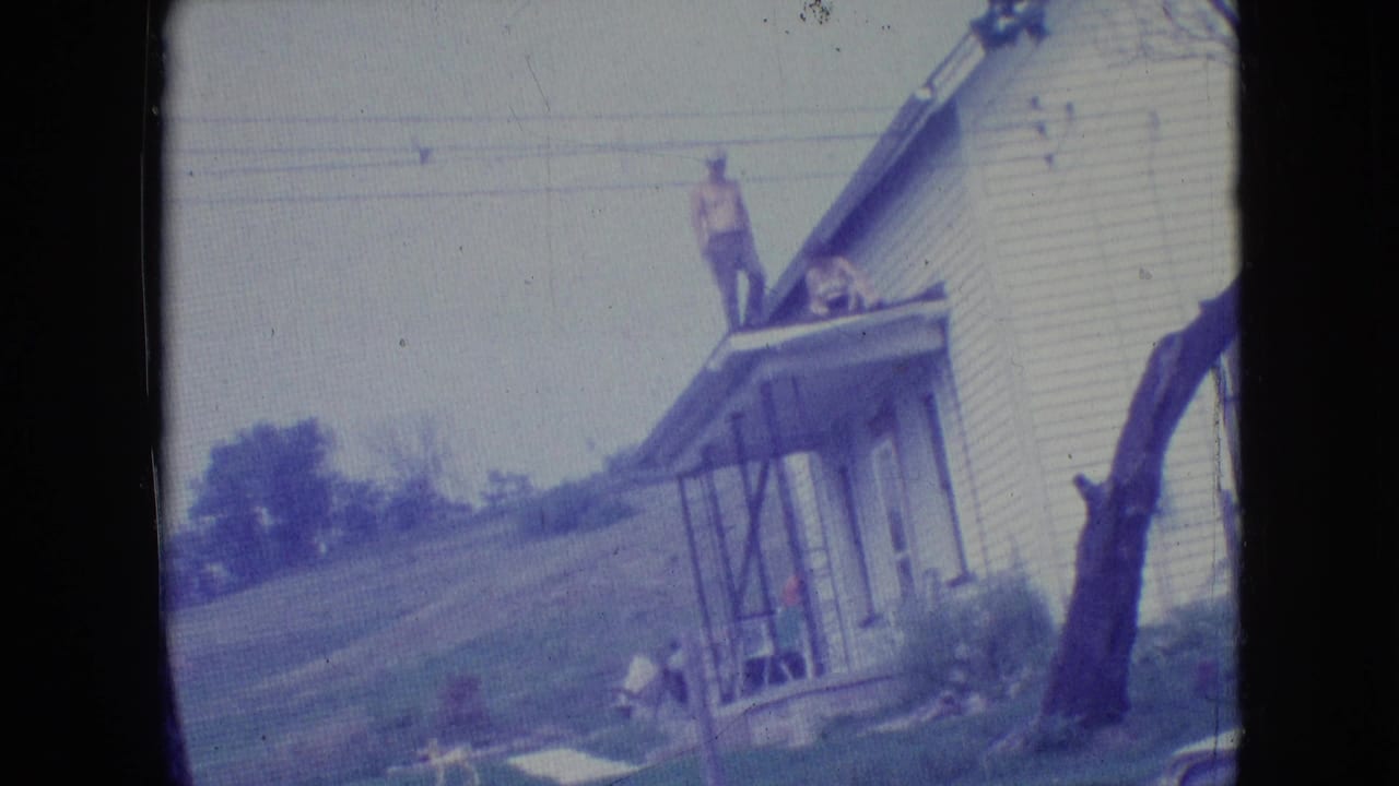 1964 Philadelphia-area 8mm home-movie frame showing a man on the roof or porch roof of a white clapboard house, with a second person near the porch and a grassy slope behind.