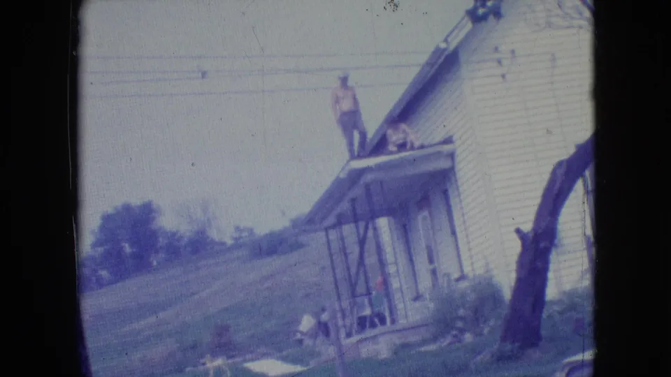1964 Philadelphia-area 8mm home-movie frame showing a man on the roof or porch roof of a white clapboard house, with a second person near the porch and a grassy slope behind.