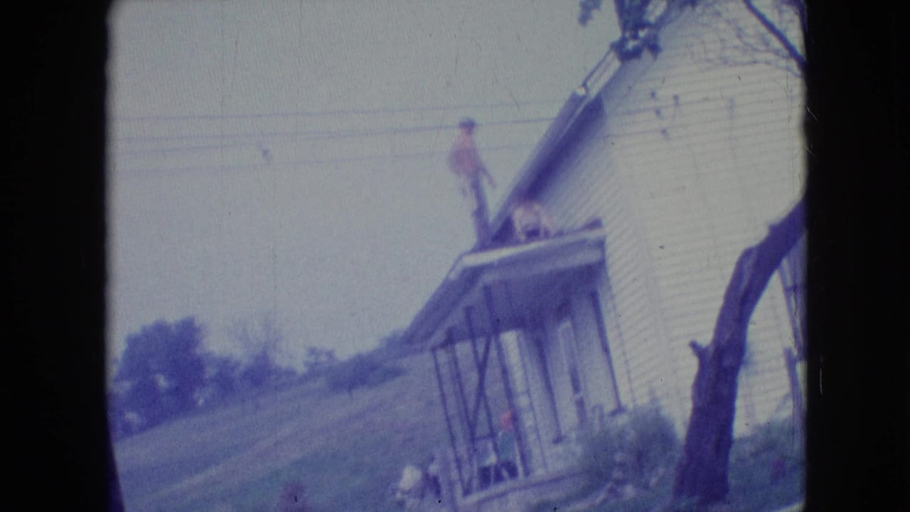 1964 Philadelphia-area 8mm frame with a man standing on the roof edge or porch roof of a white clapboard house, seen in soft faded color with another person partly visible below.
