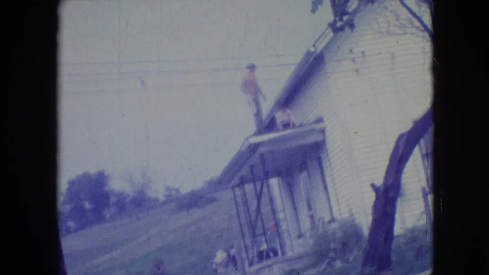 1964 Philadelphia-area 8mm frame with a man standing on the roof edge or porch roof of a white clapboard house, seen in soft faded color with another person partly visible below.