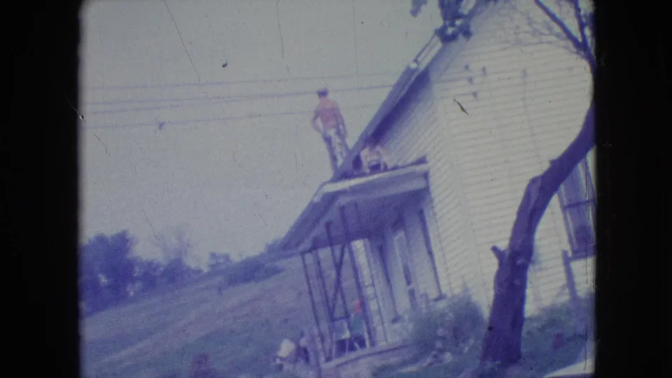 1964 Philadelphia-area home-movie frame showing a man in work clothing on the roof or porch roof of a white clapboard house, with a second person near the porch and lawn below.