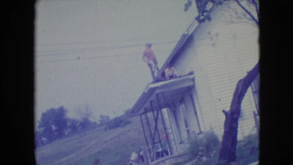 1964 Philadelphia-area 8mm frame of a man on a roof or porch roof during home repair, with a white clapboard house, front porch, tree trunk, and sloping yard visible.