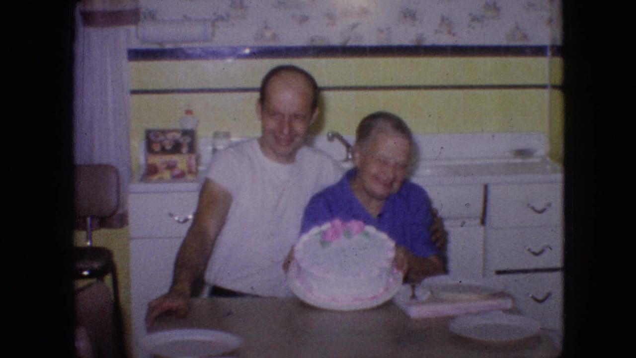In a 1964 Philadelphia home-movie frame, an older man in a white T-shirt stands beside an older woman in a blue top as she holds a decorated cake in a kitchen.