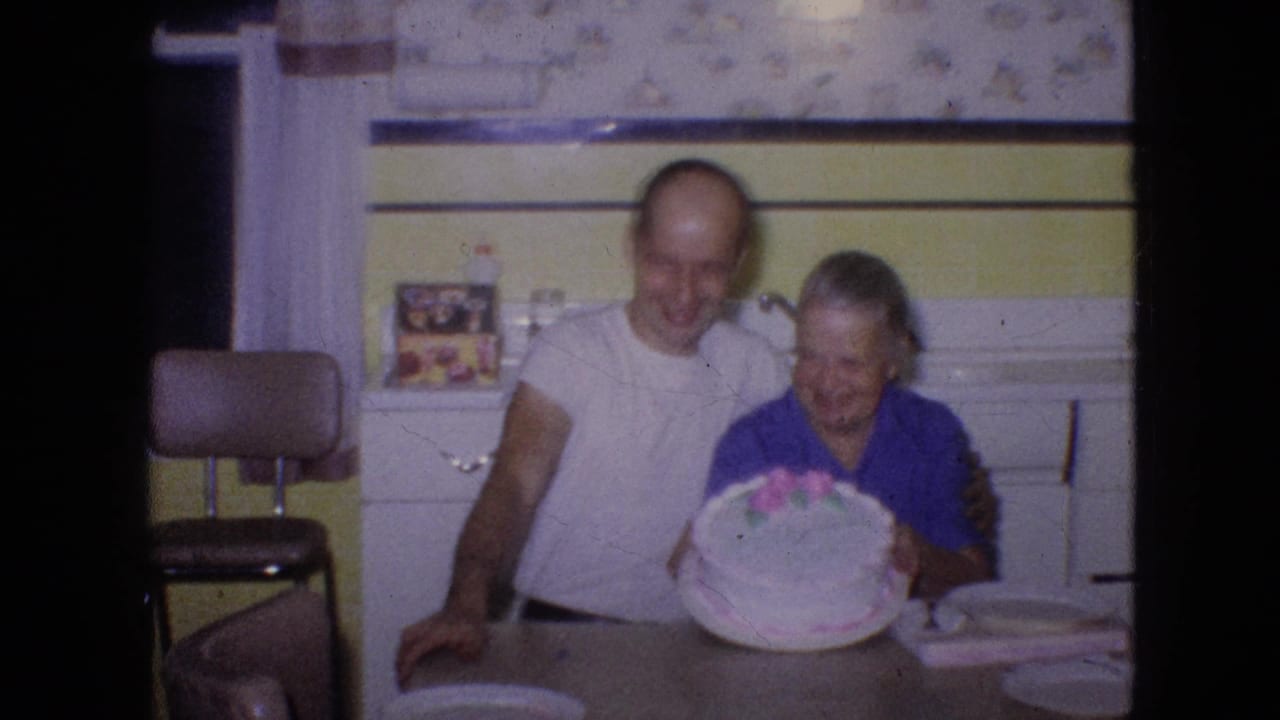The couple smiles toward the camera in a Philadelphia kitchen, with the older woman holding a round birthday cake and paper plates set on the table.