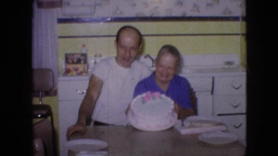 An older couple leans together behind a kitchen table, smiling as the decorated cake is held forward in this 1960s home-movie birthday scene.
