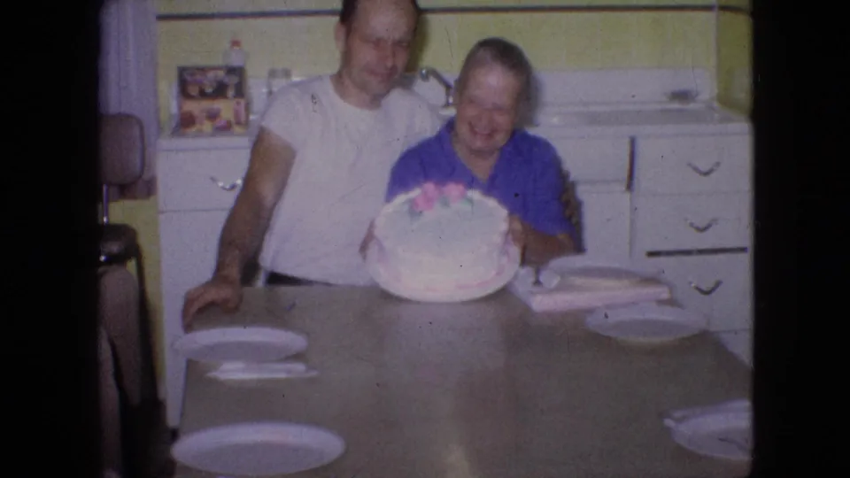 At the end of the clip, the older woman presents the round decorated cake while the older man stands close beside her in the private kitchen interior.