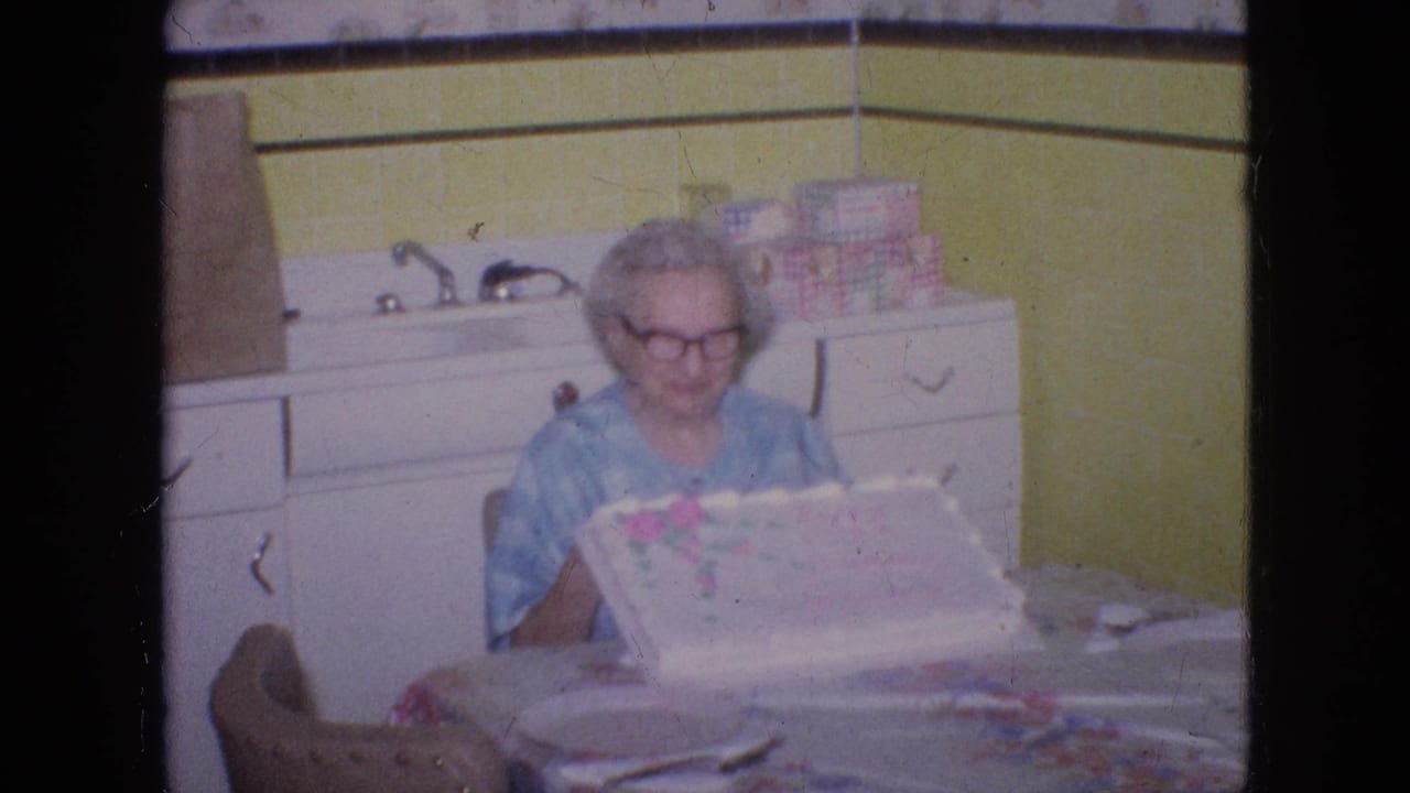 In a Philadelphia 1964 home-movie frame, an older woman in glasses and a blue patterned dress sits at a kitchen table with a decorated cake in front of yellow wall tile and white cabinetry.