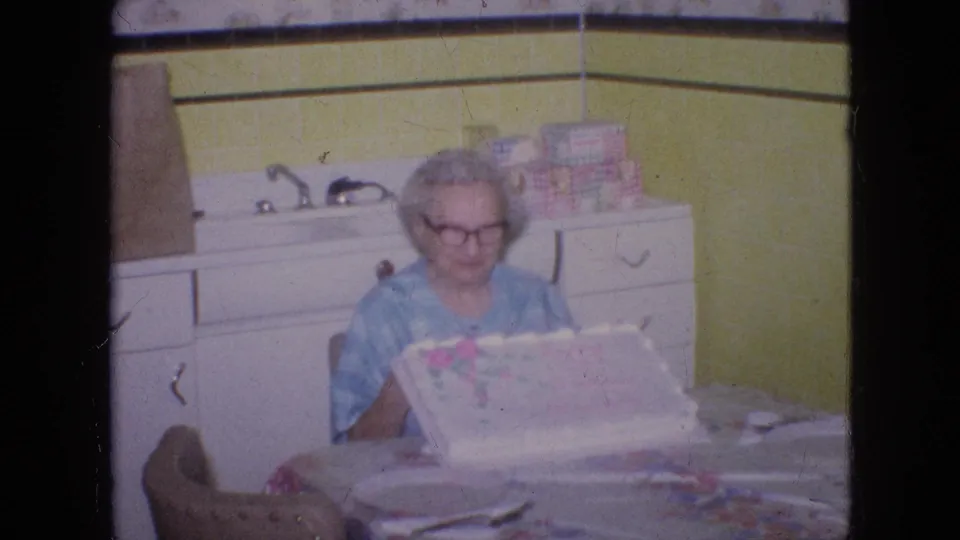 In a Philadelphia 1964 home-movie frame, an older woman in glasses and a blue patterned dress sits at a kitchen table with a decorated cake in front of yellow wall tile and white cabinetry.