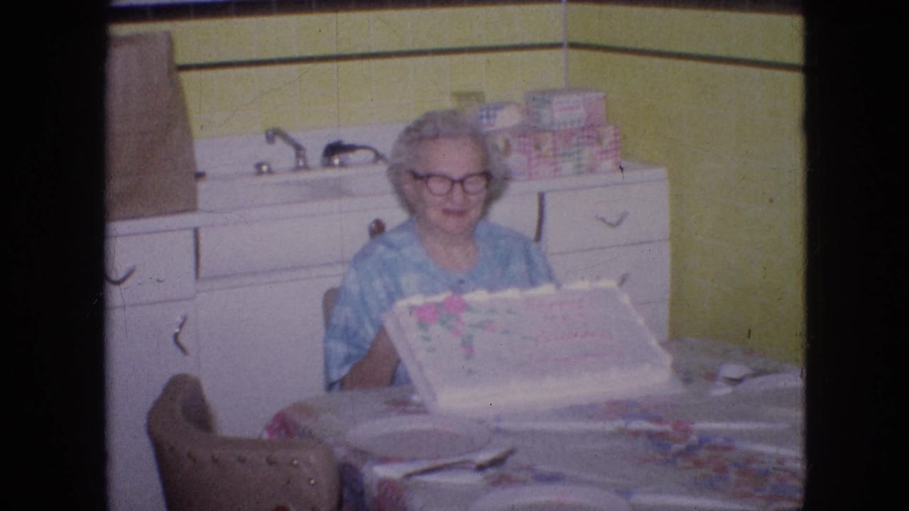 Older woman smiles while seated at a table with a decorated cake in a kitchen.
