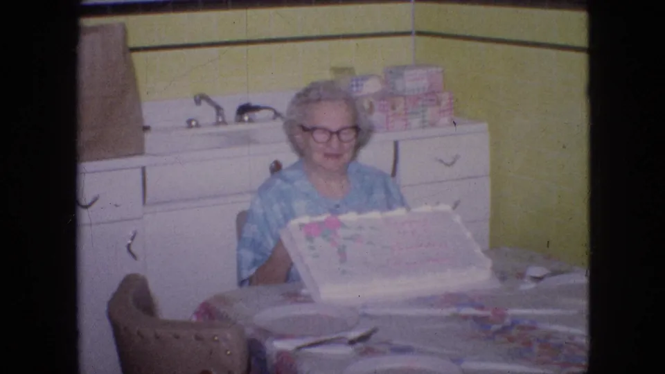 The older woman appears to smile at the table with the decorated cake, with white cabinetry, sink fixtures, yellow tile, and stacked household items visible behind her.