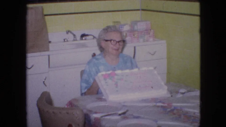 A 1960s domestic kitchen view shows the older woman seated behind the decorated cake, with plates, utensils, a floral tablecloth, yellow walls, and white cabinetry around her.