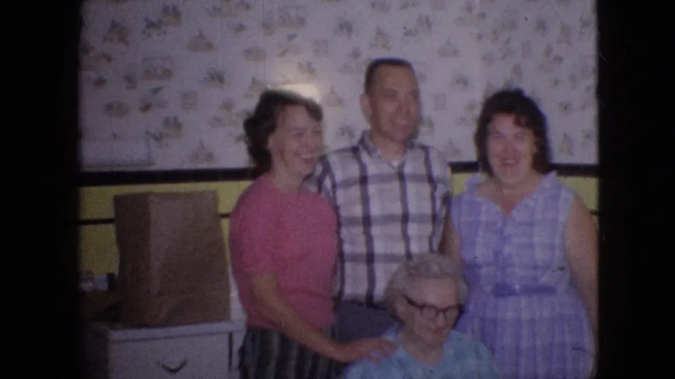 Three adults in 1960s clothing stand close together behind the seated older woman in the kitchen, suggesting a posed family moment around the celebration.