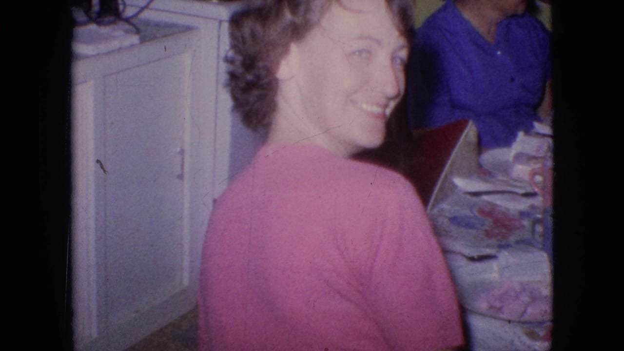 In a 1964 Philadelphia home-movie frame, a woman in a pink sweater turns from a crowded kitchen table and smiles toward the camera while other adults sit nearby.