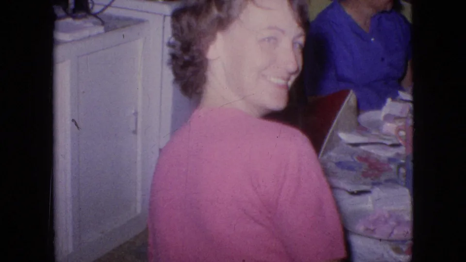In a 1964 Philadelphia home-movie frame, a woman in a pink sweater turns from a crowded kitchen table and smiles toward the camera while other adults sit nearby.