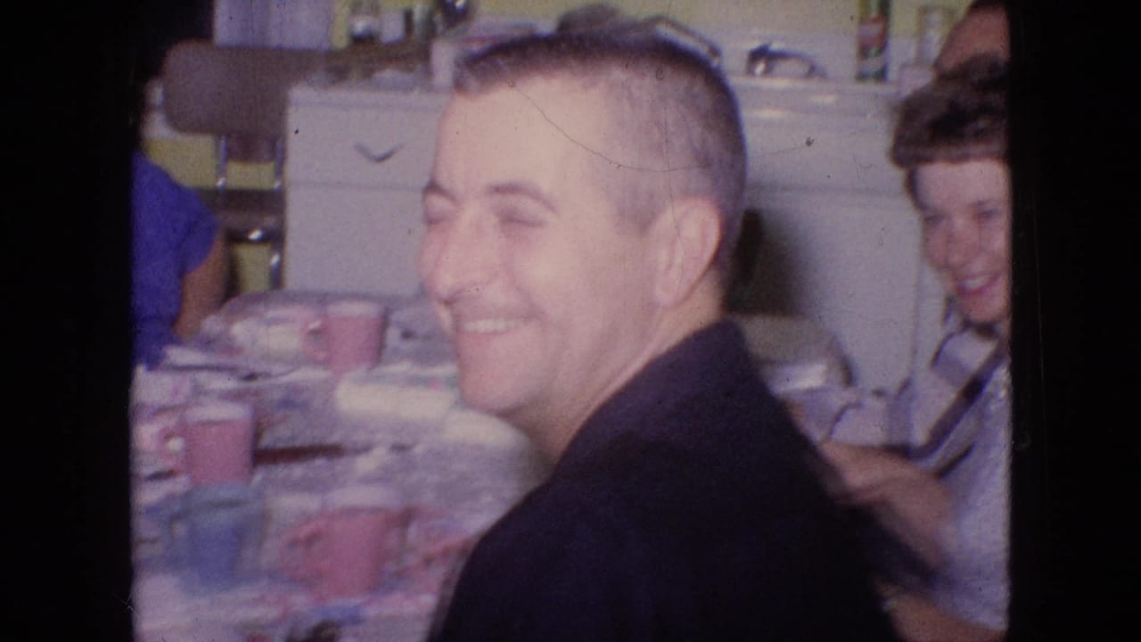 Man in a dark shirt smiles at a kitchen table with other adults nearby.