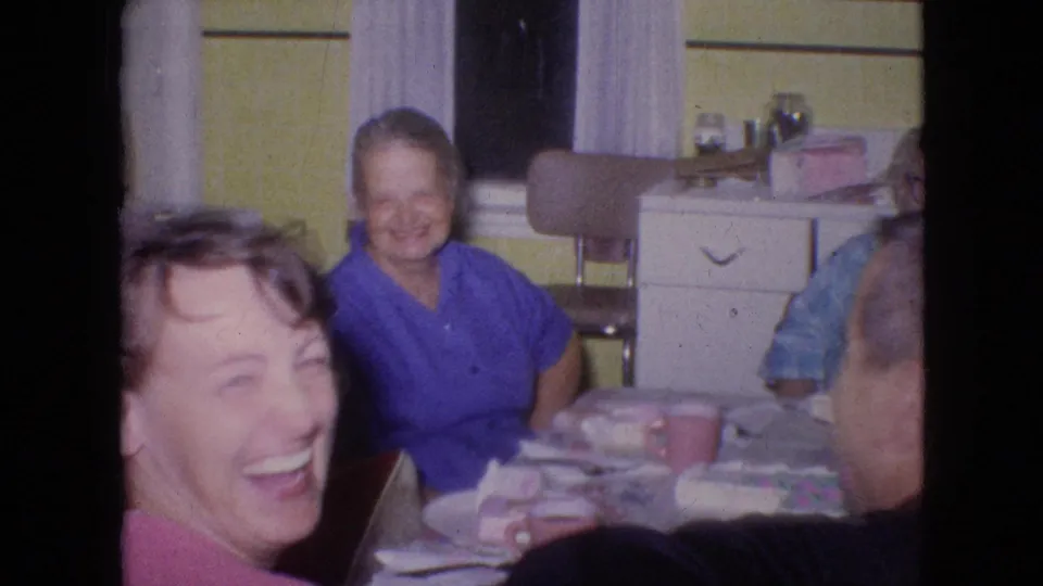 Adults laugh and sit close together around a cluttered kitchen table in a 1964 Philadelphia home-movie frame, with a woman in pink nearest the camera and another adult in blue at the back.