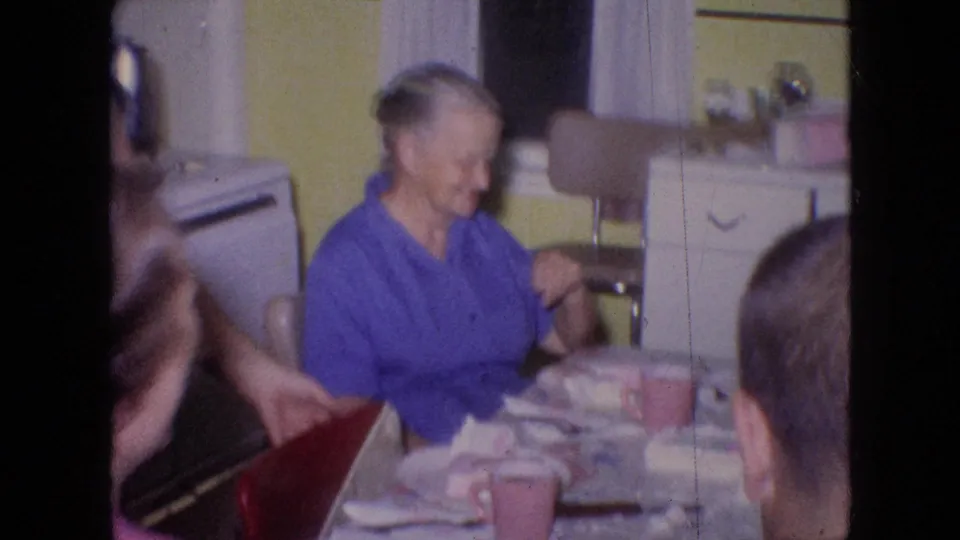 A 1964 Philadelphia home-movie frame shows seated adults continuing the kitchen-table gathering, with a person in blue near the center and tableware spread across the foreground.