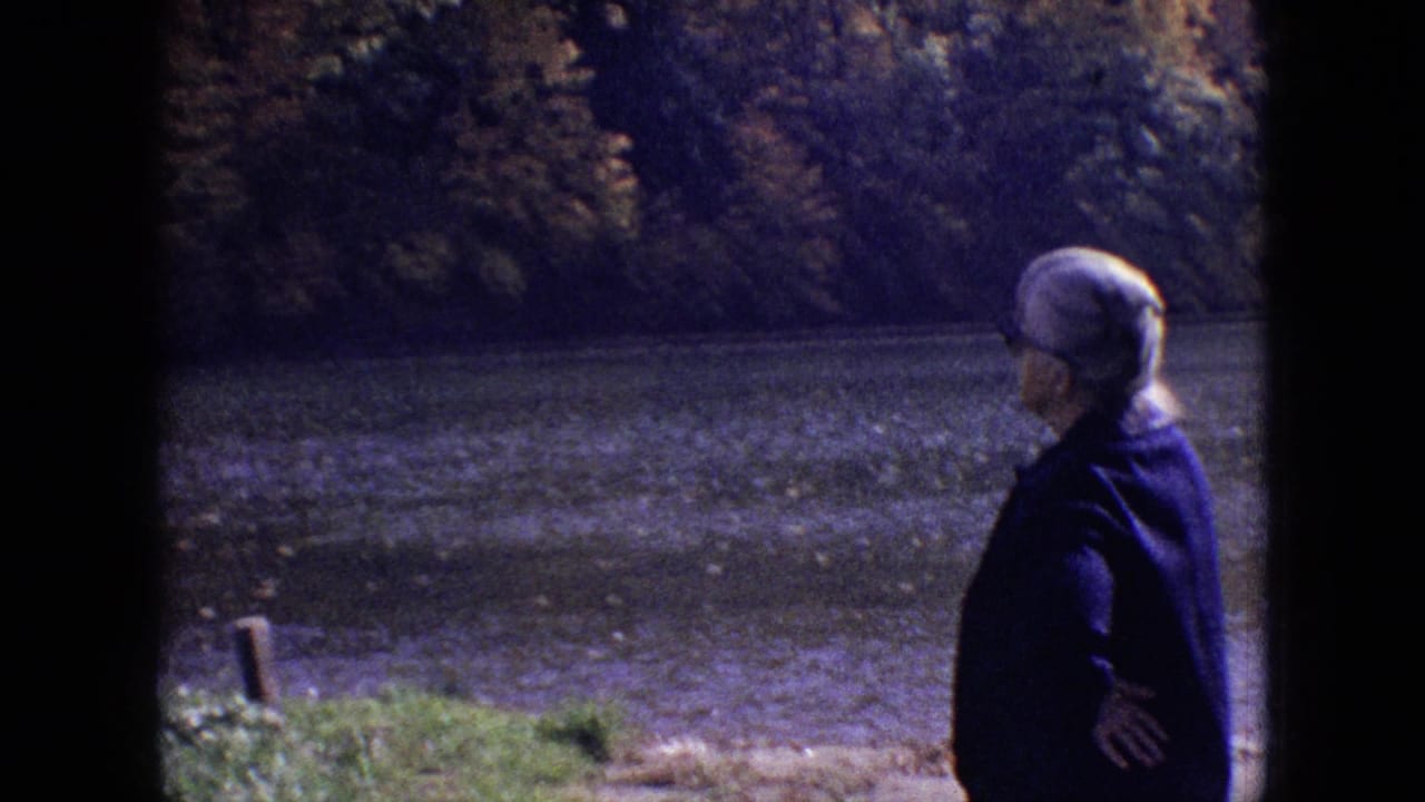 1964 Philadelphia-area home-movie frame showing an older woman in a dark coat and light head covering standing near the riverbank.