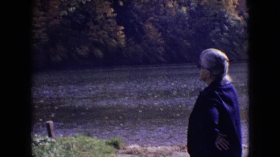 1964 Philadelphia-area home-movie frame showing an older woman in a dark coat and light head covering standing near the riverbank.