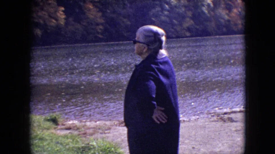 1964 Philadelphia home-movie frame with an older woman in profile near a calm river, framed by visible 8mm borders and autumn trees.