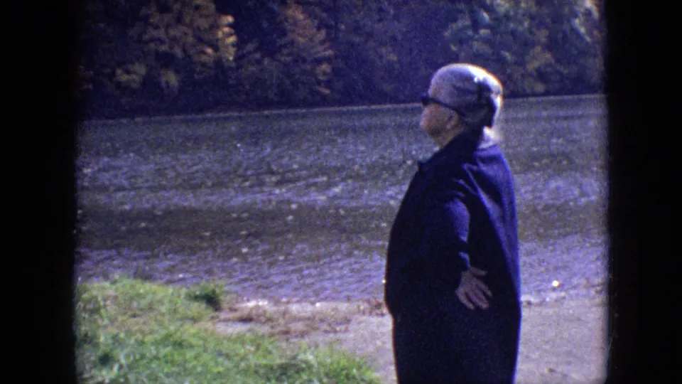1964 Philadelphia-area 8mm frame showing an older woman paused at the river's edge with trees and water filling the background.