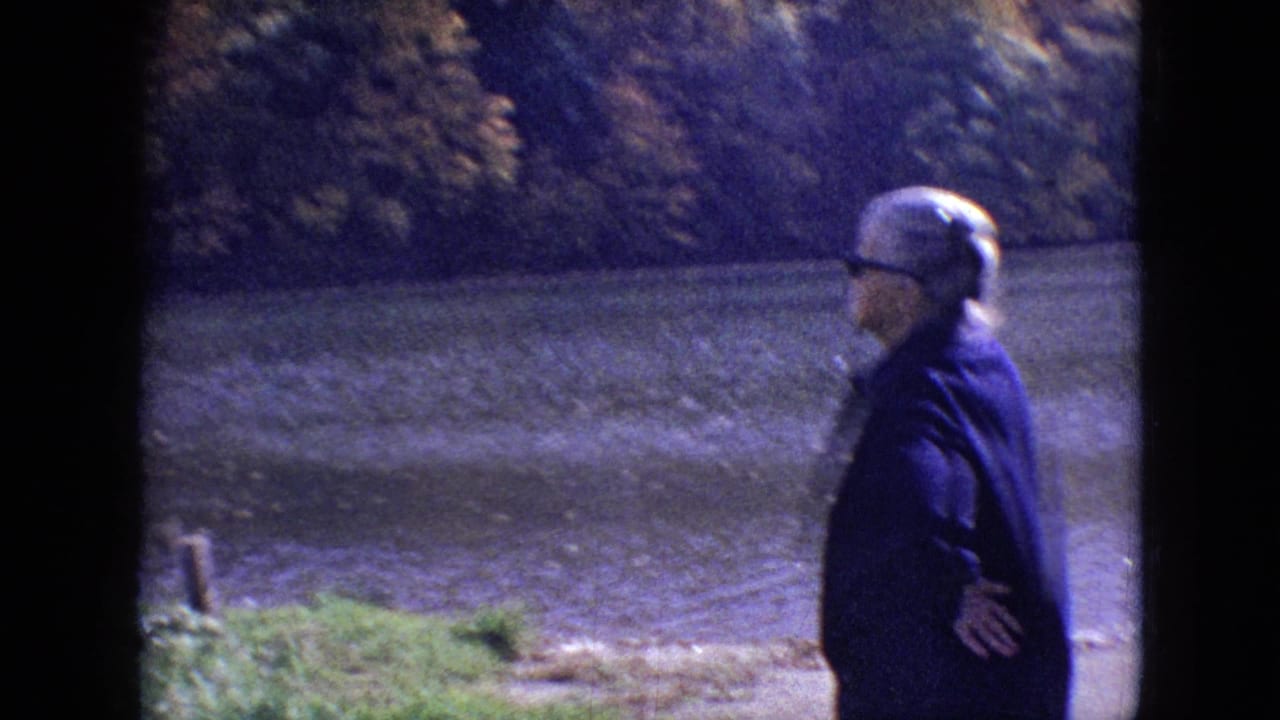 In 1964 Philadelphia home-movie footage, a woman in a dark coat and sunglasses stands near rippling lake water with fall-colored trees across the shore.