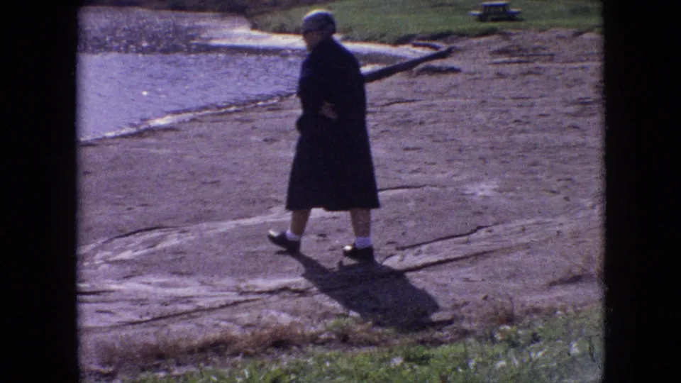 1964 8mm home-movie frame from Philadelphia showing a person in a dark coat and hat walking along the sandy shoreline beside the lake, with a long shadow on the ground.