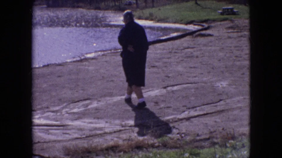In 1964 Philadelphia home-movie footage, a woman in a dark coat and hat walks away from the camera along a receding riverbank beside calm water and scattered logs.