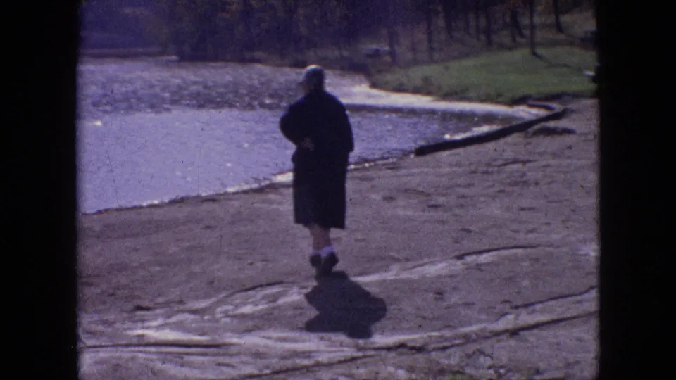 The woman continues walking away near the water's edge, with a curving shoreline, grass, and logs visible in the background of the 1964 Philadelphia footage.