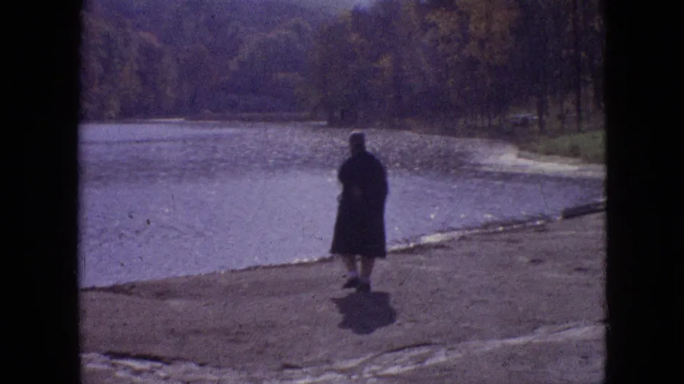 The final frame places the woman farther from the camera on the exposed shoreline, silhouetted against the calm water and wooded bank in the 1964 Philadelphia clip.