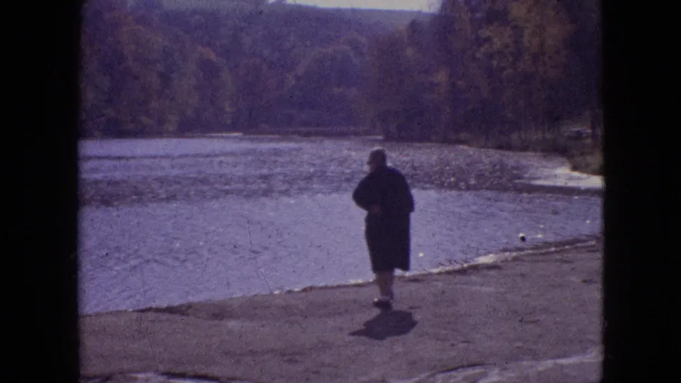 1964 Philadelphia home-movie frame showing a distant adult visitor walking near a water edge with trees beyond, before the marine mammal pool views.