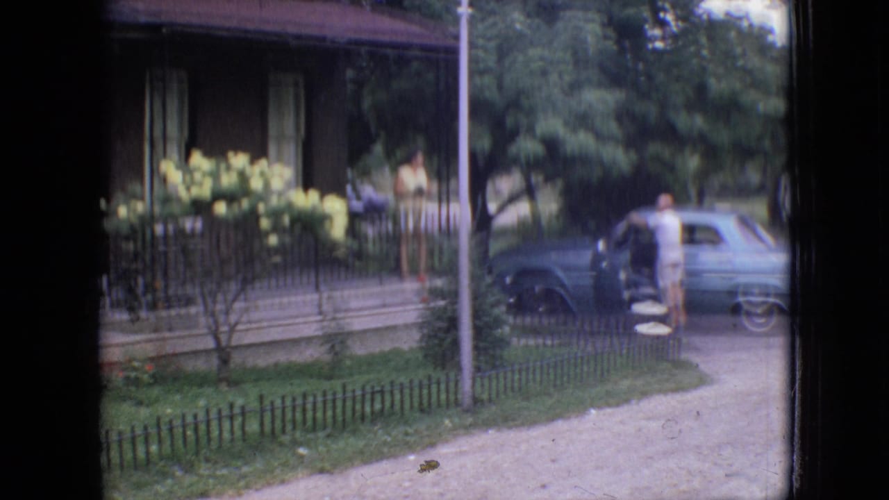 1964 Philadelphia home-movie frame showing a light blue vintage car near a residential porch with yellow flowers, trees, grass, and a person standing by the open car door.