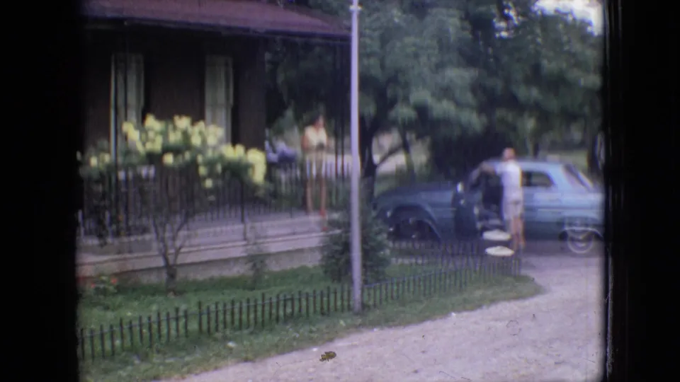 1964 Philadelphia home-movie frame showing a light blue vintage car near a residential porch with yellow flowers, trees, grass, and a person standing by the open car door.