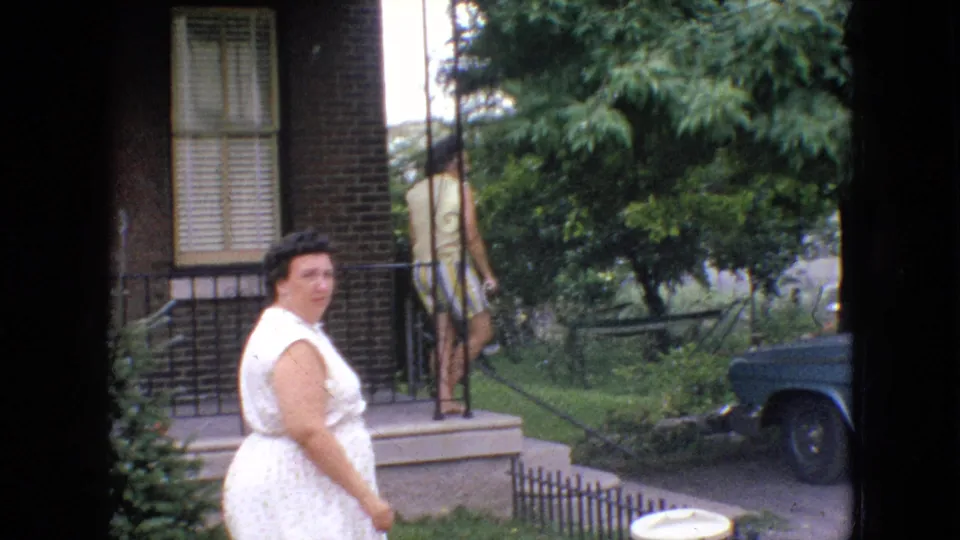 1964 Philadelphia home-movie frame showing an adult woman in a white dress beside a brick residential porch, with metal railings, trees, lawn, and part of a parked car visible.