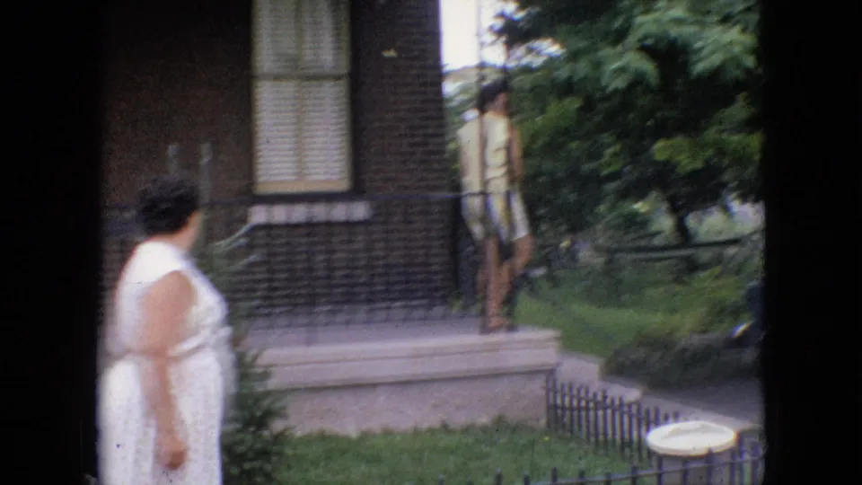 1964 Philadelphia home-movie frame showing one adult woman in a white dress approaching porch steps while another adult woman in yellow clothing stands near the railing.