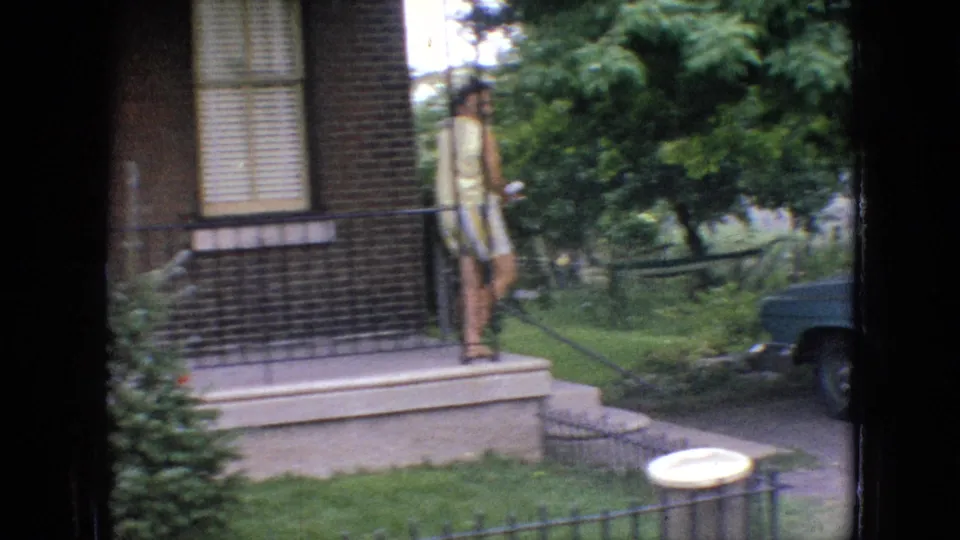 1964 Philadelphia home-movie frame showing an adult woman in yellow clothing on residential porch steps, with black railings, brick facade, trees, lawn, and a partial blue car at the edge of frame.
