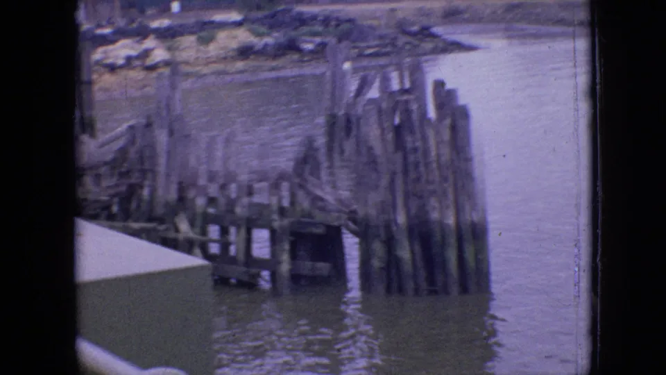 Closer 1964 Philadelphia waterfront frame showing broken dock framing, vertical pilings, and water around the structure, with visible 8mm grain and fading.